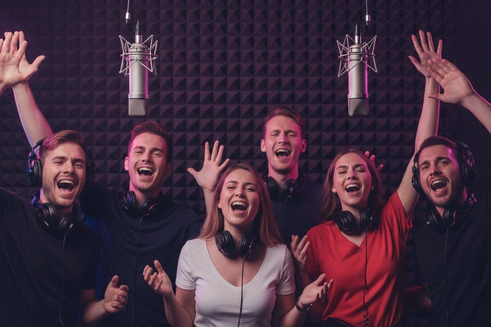 Six amis chantent ensemble devant deux micros de studio, bras levés, casques autour du cou